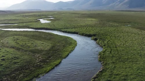 Aerial view of winding river peacefully meanders through lush green fields. Mountains in the distanc