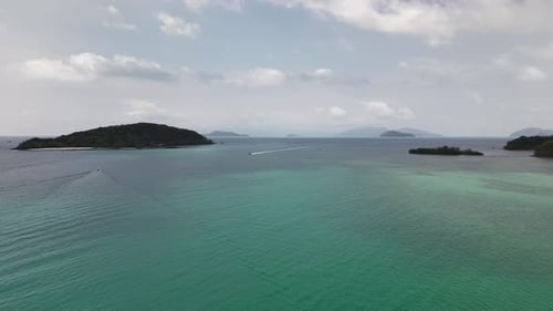 Drone glides above clear turquoise sea near tropical island as boats speed across gentle waves...