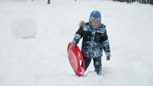 Slow motion of a happy smiling boy walking up the snowy hill with his plastic sleds at snowfall