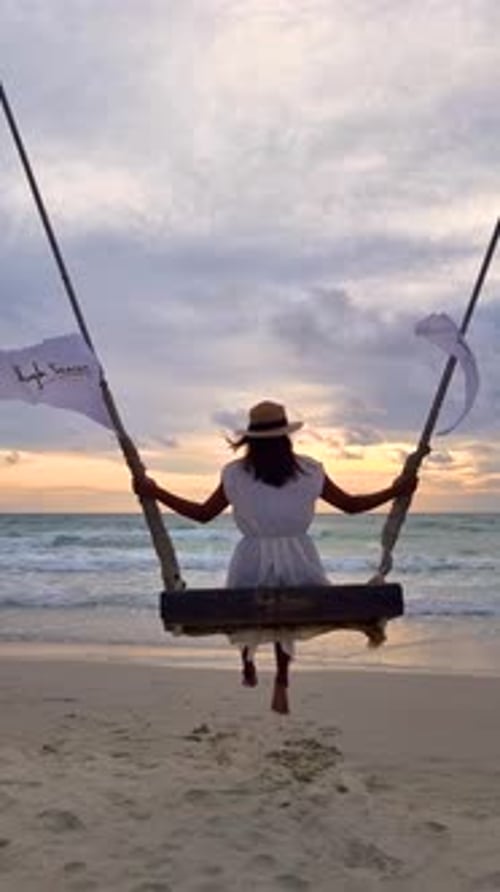 Asian Woman Watching Sunset on the Beach at a Swing Under a Palm Tree