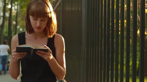 Young woman reads a interesting book and walking near iron fence at the city park
