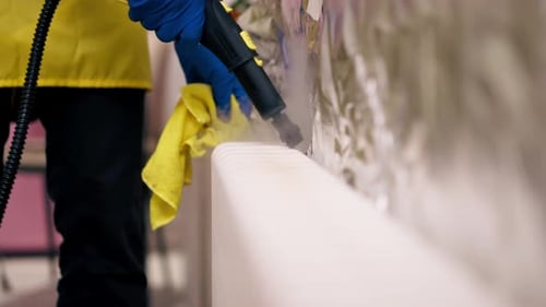 Close Up in an Apartment Cleaner in a Yellow Apron Washes a White Radiator with a Steam Generator