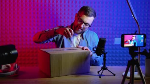 Young Smiling Blogger Unpacking Box with Present Sitting at Home Studio