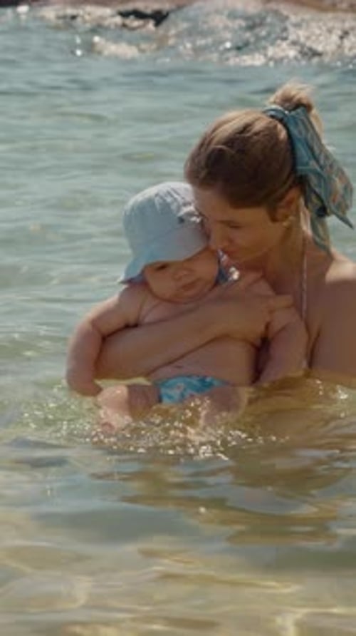 Vertical video Mother embracing her baby in shallow water at the beach