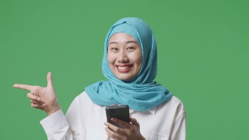 Close Up Of Asian Muslim Woman Smiling And Using Smartphone Then Pointing To Side On Green Screen