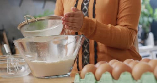 Video of midsection of biracial woman baking in kitchen at home, sieving flour into mixing bowl