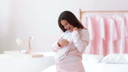 Pregnant Woman Holding White Baby Onesie in Bedroom