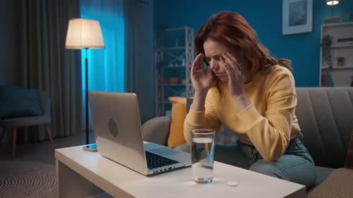 A Woman Suffers From a Headache Attack While Working on a Laptop While Sitting on a Sofa in the
