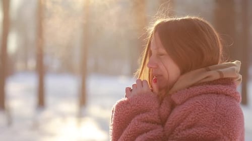 Beautiful young woman trying to warm her frozen hands with a breath in winter park. Girl with frosty