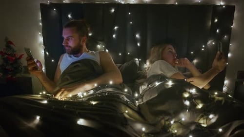 Couple in Bed Looking at Phones, String Lights