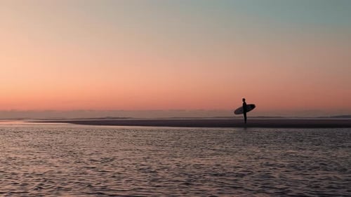 Young woman surfer holding a surfboard and looking at golden sunset / sunrise.