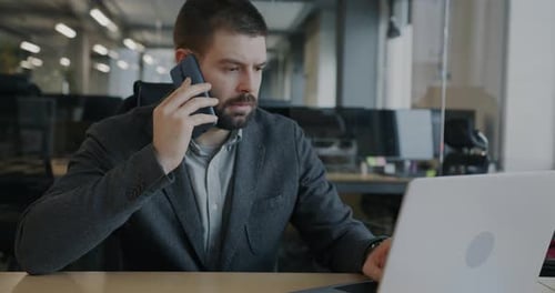 Serious Businessman Talking on Mobile Phone and Yuping with Laptop Working in Glass Wall Office