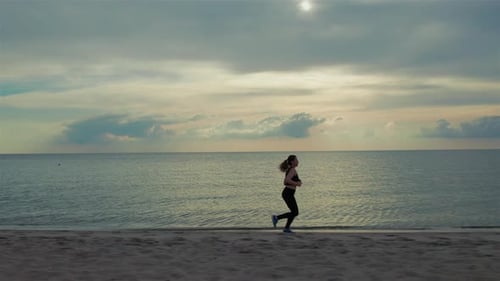 A young woman runs along the beach in sporty clothing, her hair blowing in the wind as she pushes he
