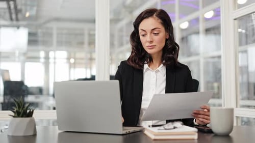 Businesswoman Working at Her Desk in Office Checking Documents and Using Laptop