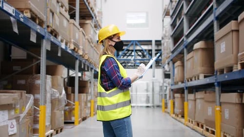 Young female warehouse worker in protective gear checks stock during pandemic