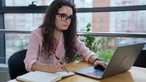 Businesswoman sitting at table in a spacious office, working with laptop and taking notes.