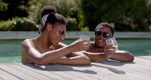 African American couple glancing, turning toward each other at pool edge, clinking glasses toasting