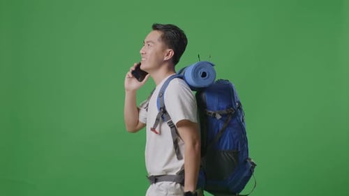 Side View Of Asian Male Hiker Talking On Smartphone While Walking On Green Screen Background