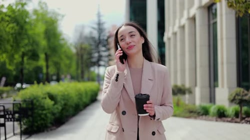 Businesswoman Talking on Phone and Holding Coffee Cup While Walking Along Street