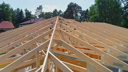 Aerial view over the wooden beams of a roof of a house in unfinished construction on green neighbour