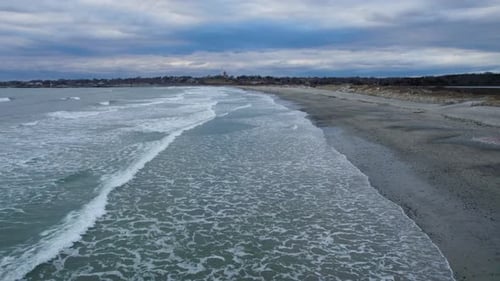 Aerial of ocean waves crashing on a beach on a cloudy day. Drone of sea water slowly crashing on the