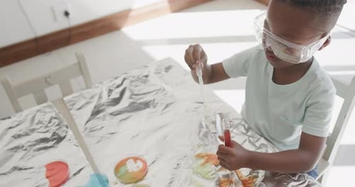 Niño afroamericano sentado en la mesa con tubos de ensayo con líquido, en cámara lenta