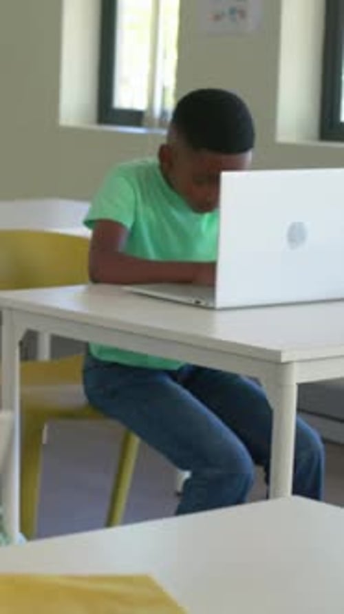 Vertical video: Camera panning teen in green tee typing on white laptop in classroom doing homework