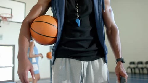 Holding basketball, coach with whistle standing in school gym, players practicing