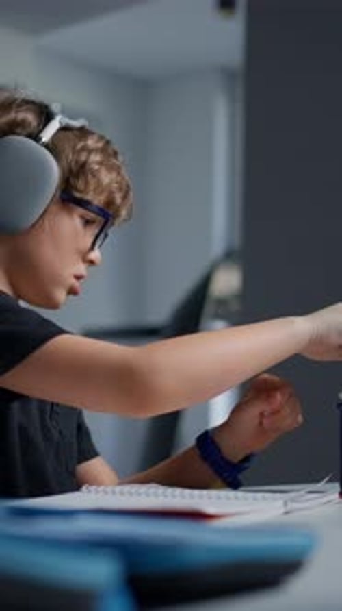Young Boy with Glasses and Headphones Focused on Studying and Writing in a Notebook at a Desk in a