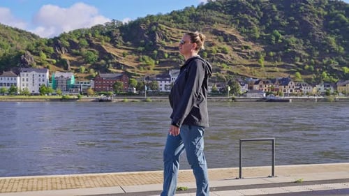 Woman walking along the banks of the Rhine River in the town of Sankt Goar, with the town St. Goar
