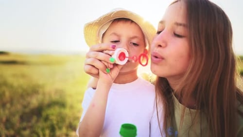 Teenage Sister Stands and Smiles Next to Little Brother Who Blows and Gets Soap Bubbles in Outdoor