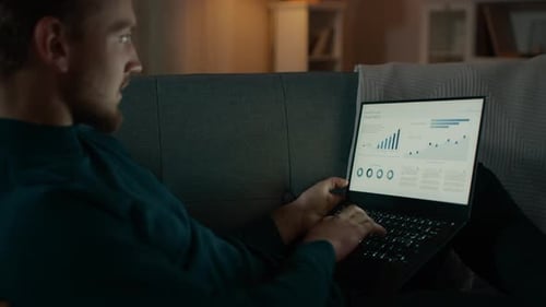 In the Evening Handsome Young Man Relaxes at Home Laying on a Couch in the Living Room Works on a L