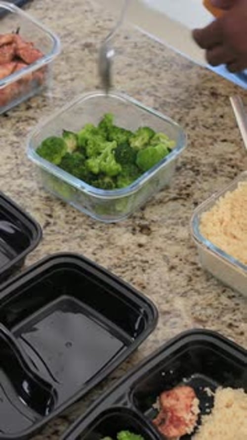 Vidéo verticale : Soulevant une cuillère de brocoli, un homme au dessus orange emballe du poulet dans des plateaux à la maison