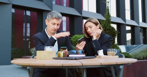 Middle-aged businessman working on laptop at an outdoor cafe while having a business call on phone.