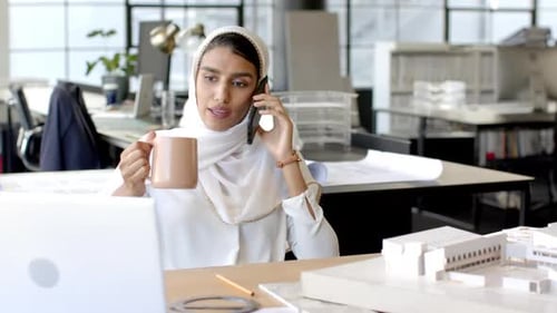 Talking on smartphone, woman working on laptop in modern office environment