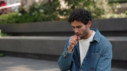Portrait of Young Hispanic Man with Beard Coughs Covering Mouth By Hand in Park
