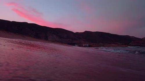 Camera resting on shore sand of the beach while waves break over it in pink sunset.
