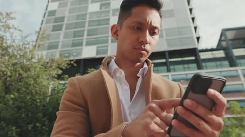 Close up, young businessman is sitting on bench in the street and typing on mobile phone