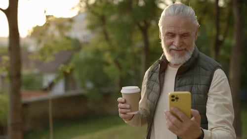 Cheerful Serion Man Using Smartphone and Carrying Cup of Coffee While Walking in the Park