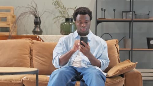 African Man Text Messaging on Phone while Sitting on Sofa at Home