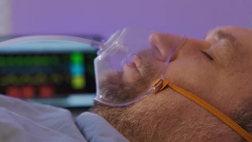 Side View of Mature Caucasian Man Wearing Oxygen Mask Lying in Bed in Room of Clinic Close Up