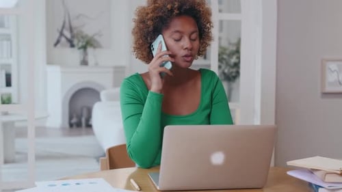 African American Woman Talking on Phone Sits at Table with Laptop in Home Office