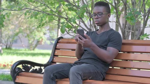 Junger Afrikaner benutzt Smartphone, während er draußen auf einer Bank sitzt