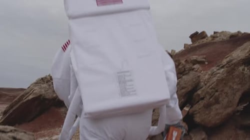 A person wearing an astronaut suit walks across a rocky terrain