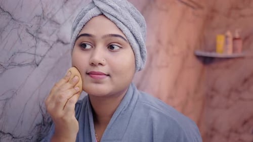 Closeup portrait of a woman blending foundation with a beauty blender in modern bathroom.
