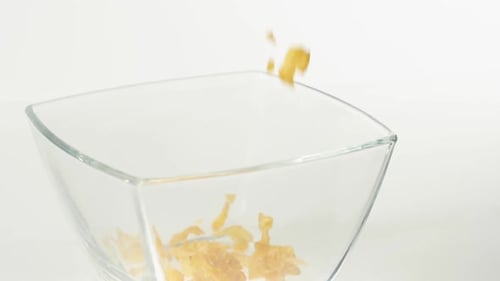 Yellow cornflakes fall in a transparent square bowl on a white background. Diet, detox, vegetarian
