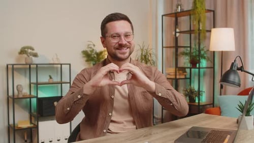 Happy Caucasian Young Man Makes Symbol of Love Showing Heart Sign to Camera at Home Office Desk