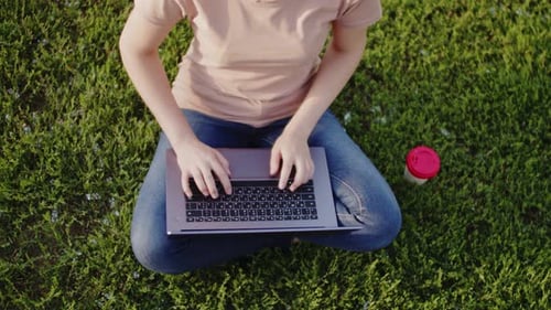 Frau sitzt mit Laptop auf Gras im Park und tippt eine Nachricht, daneben steht eine Kaffeetasse