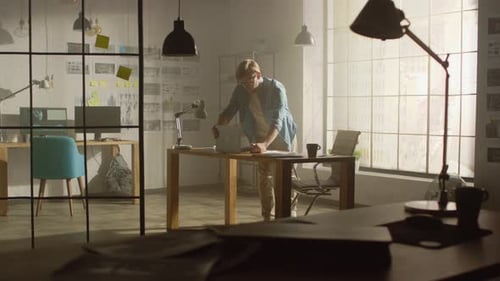 Professional Creative Designer Holds Laptop, Walks around His Office Checking Product Drawings and