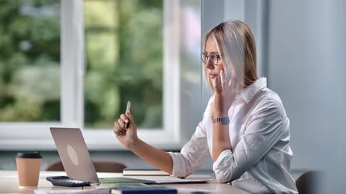 Modern Blonde Woman Boss Working Laptop Routine Paperwork Talking Smartphone at Desk Office Window